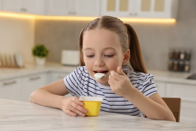 Photo of Cute little girl eating tasty yogurt at white marble table in kitchen