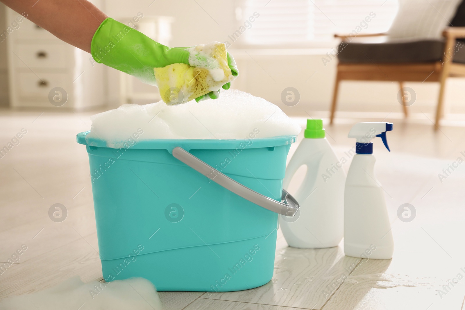 Photo of Woman holding sponge with foam over bucket indoors, closeup. Cleaning supplies