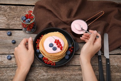 Woman putting natural yogurt onto tasty pancakes with blueberries and red currants at wooden table, top view