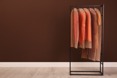 Photo of Garment bags with clothes on rack near brown wall. Space for text