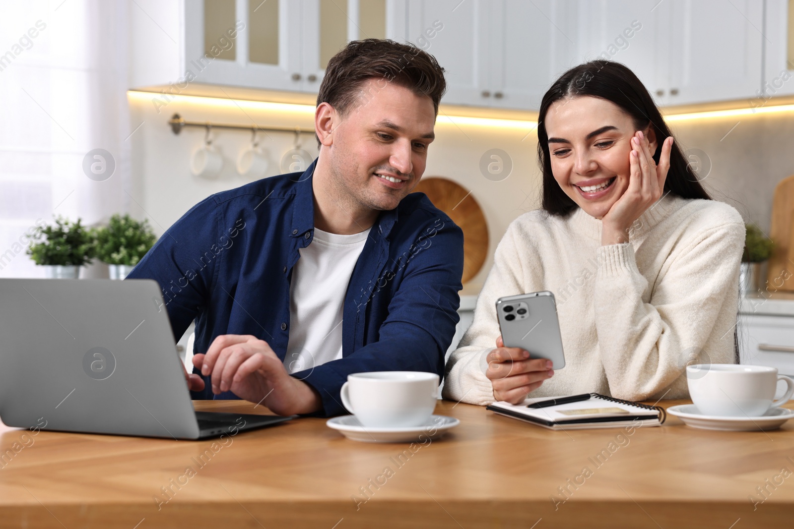 Photo of Happy couple with gadgets shopping online at wooden table in kitchen