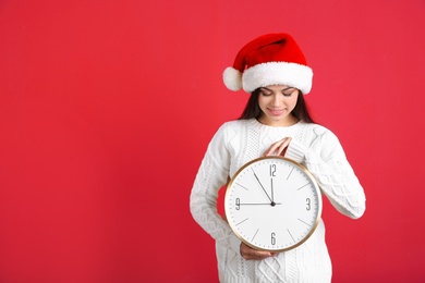 Young beautiful woman in Santa hat holding big clock on color background. Christmas celebration