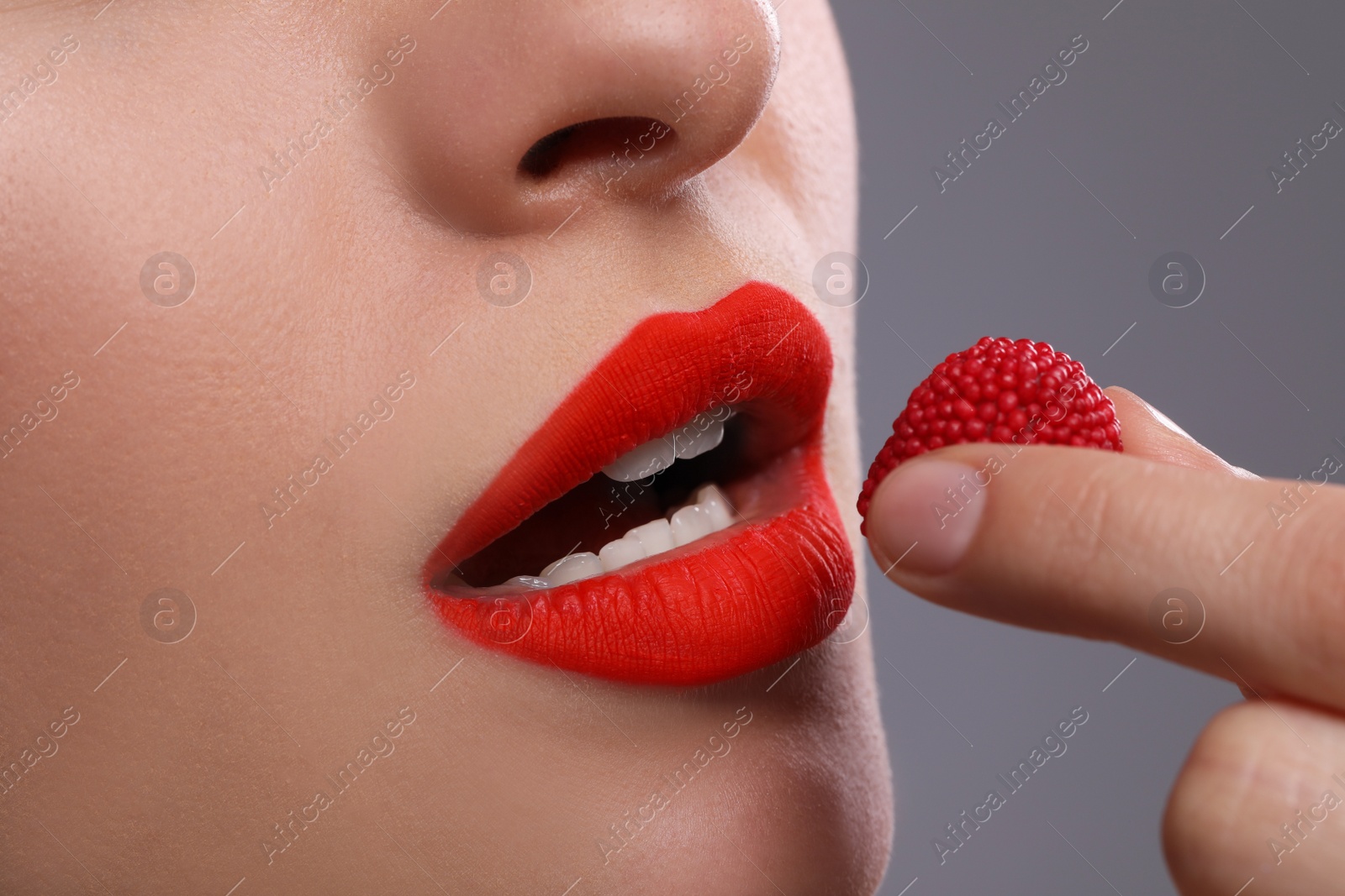 Photo of Young woman with beautiful red lips makeup eating candy on grey background, closeup