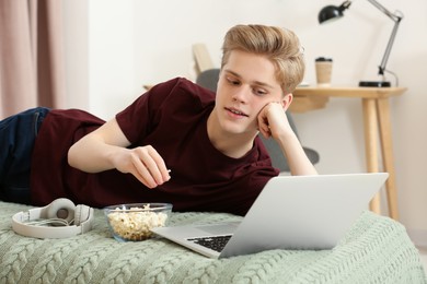 Teenage boy with popcorn using laptop on bed at home