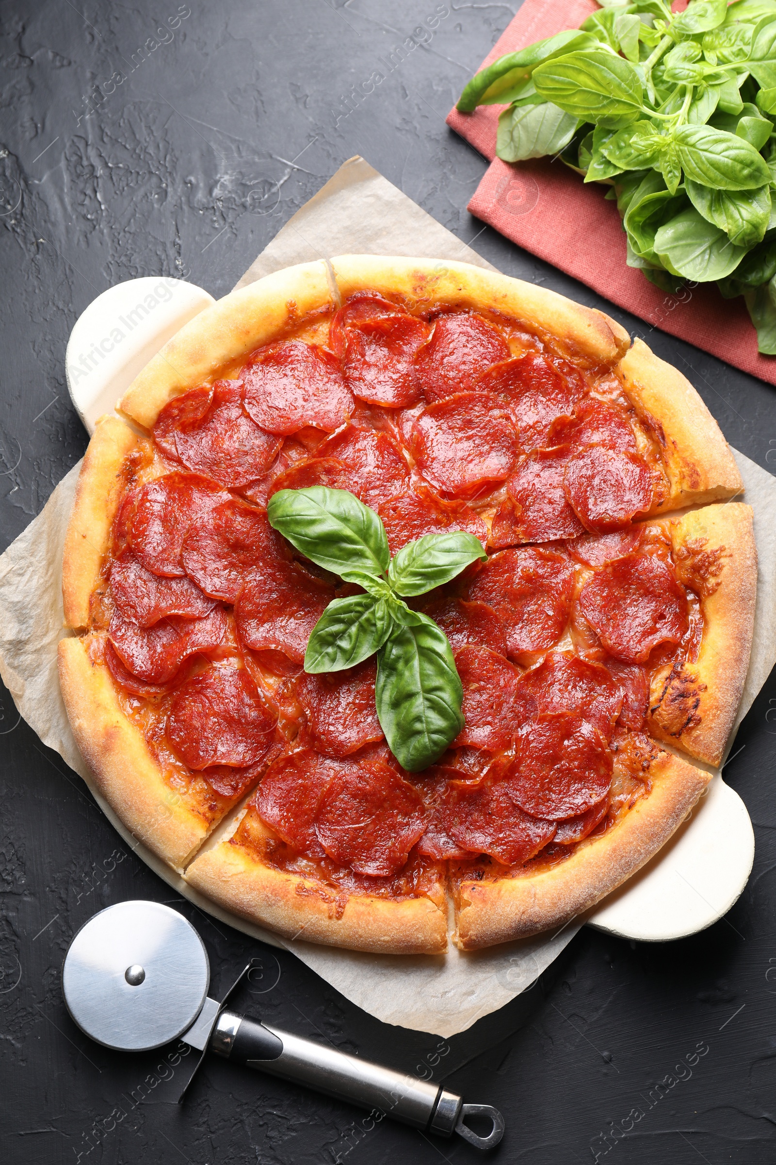 Photo of Tasty pepperoni pizza, cutter and basil on black textured table, flat lay