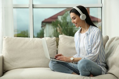 Young woman with laptop and headphones on sofa at home, space for text
