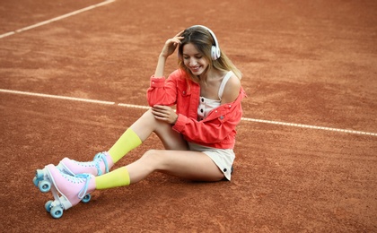 Happy stylish young woman with vintage roller skates and headphones sitting on tennis court