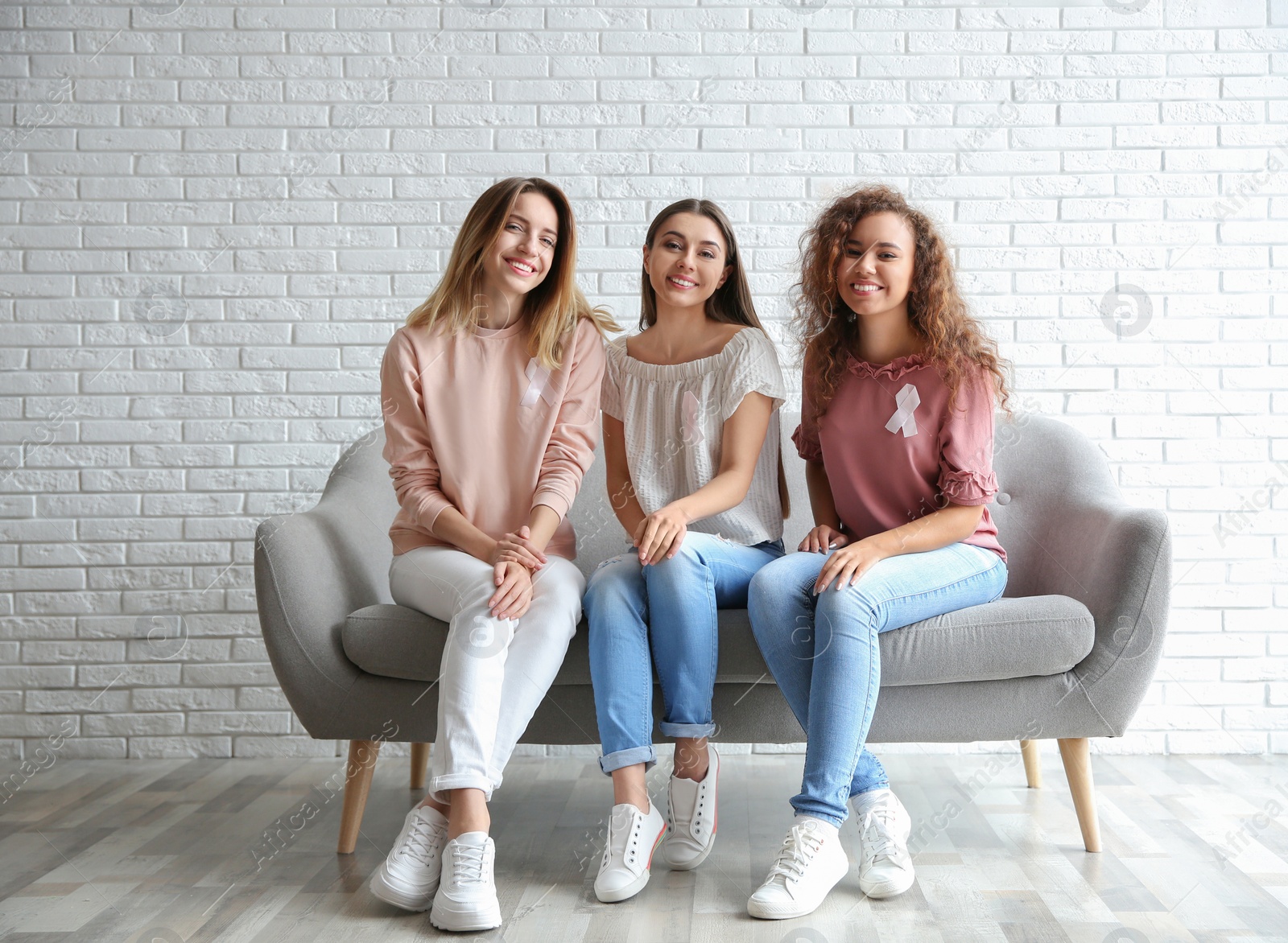 Photo of Women with silk ribbons sitting on sofa against brick wall. Breast cancer awareness concept