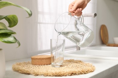Photo of Woman pouring water into glass from jug on countertop in kitchen, closeup