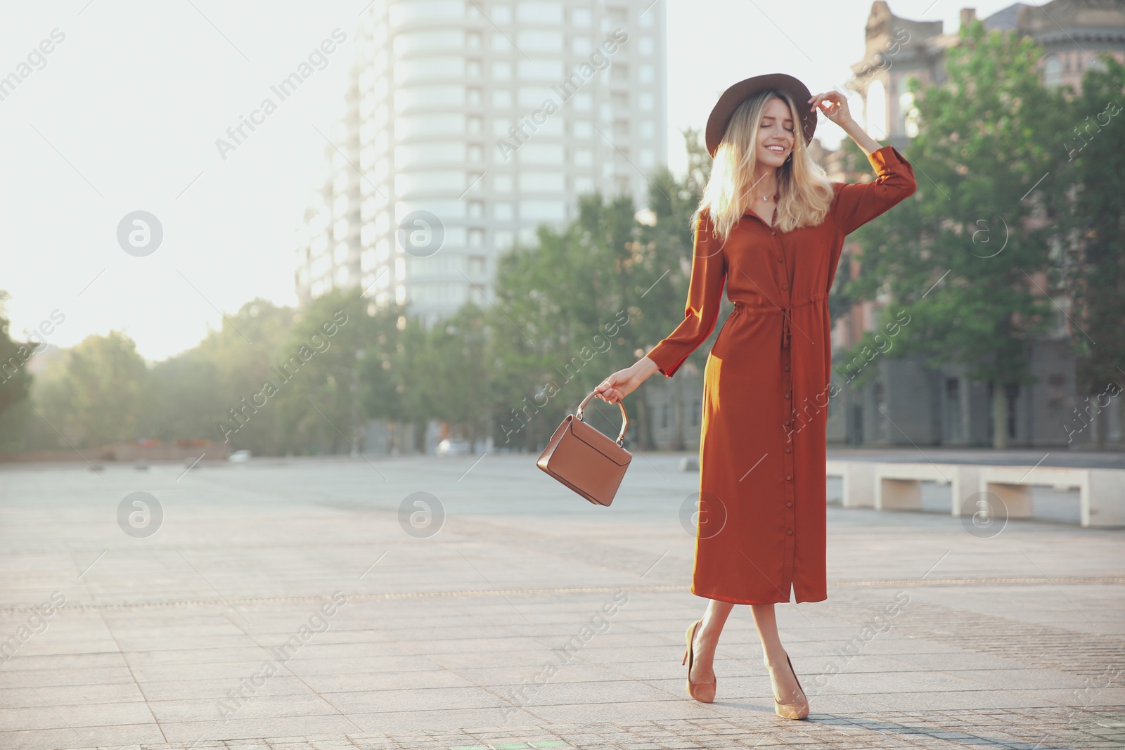 Photo of Beautiful young woman in stylish red dress and hat with handbag on city street