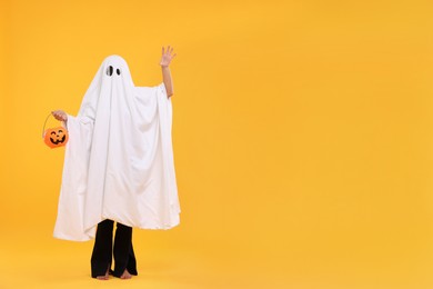 Photo of Woman in white ghost costume holding pumpkin bucket on yellow background, space for text. Halloween celebration