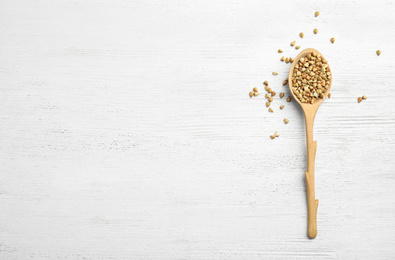 Photo of Uncooked green buckwheat grains in spoon on white wooden table, top view. Space for text