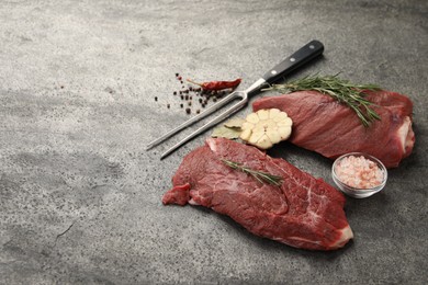 Photo of Piece of raw beef meat, spices, rosemary and fork on grey table, space for text