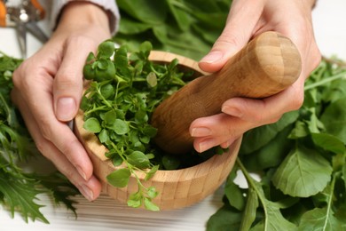 Woman grinding fresh green herbs in mortar at white wooden table, closeup