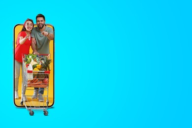 Grocery shopping via internet. Happy couple with shopping cart full of products walking out of huge smartphone on light blue background, space for text