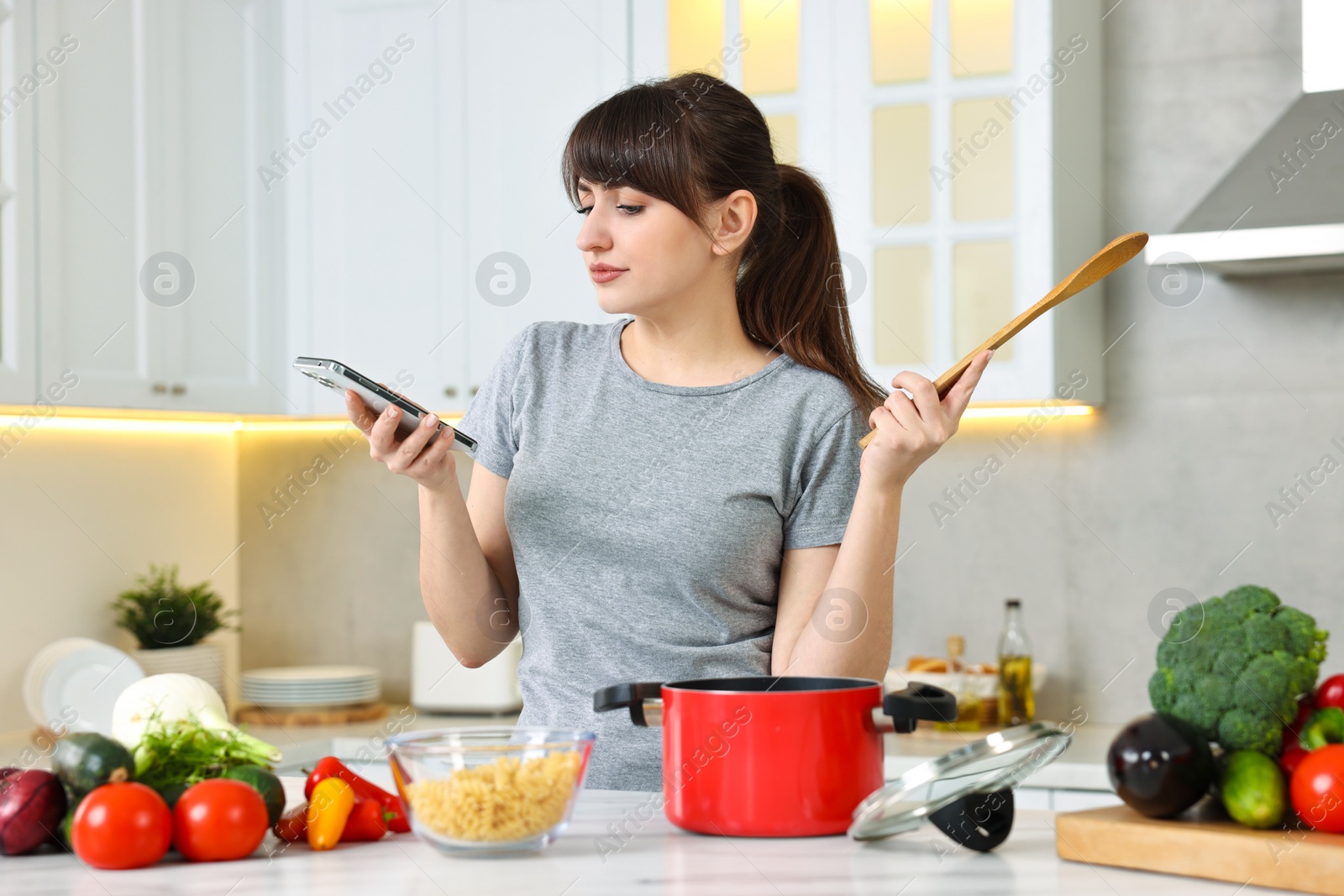 Photo of Young housewife with spoon using smartphone while cooking at white marble table in kitchen