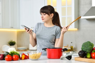 Young housewife with spoon using smartphone while cooking at white marble table in kitchen