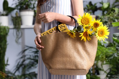 Woman holding beach bag with magazine and beautiful bouquet of sunflowers indoors, closeup