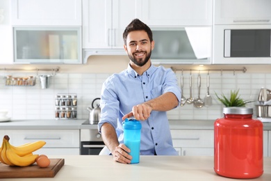 Photo of Young man opening bottle of protein shake at table with ingredients in kitchen