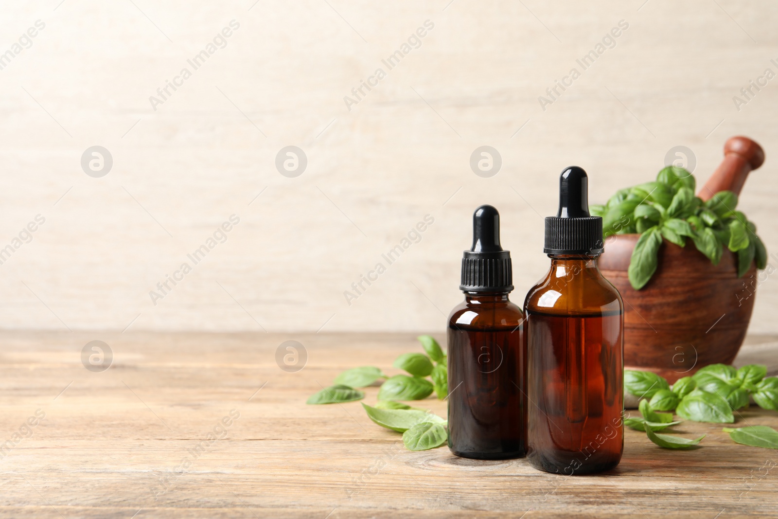 Photo of Bottles of basil essential oil near mortar with leaves and pestle on wooden table. Space for text