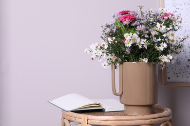 Stylish ceramic vase with beautiful flowers and open book on wicker table near light wall. Space for text