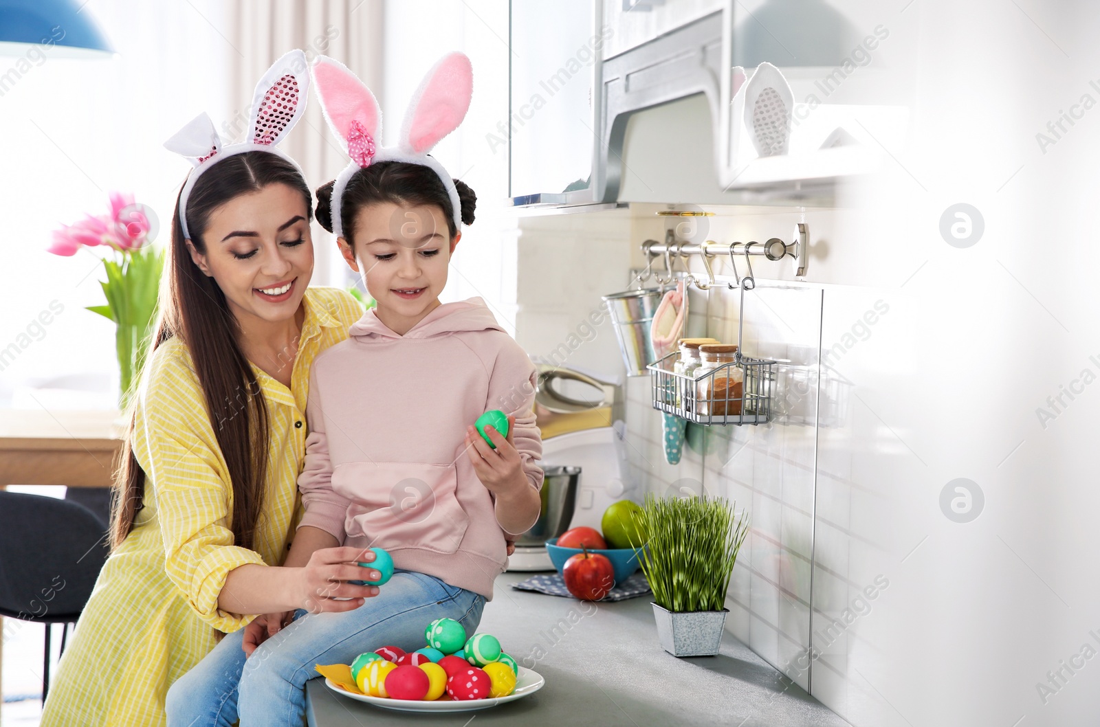 Photo of Mother and daughter with bunny ears headbands and painted Easter eggs in kitchen, space for text