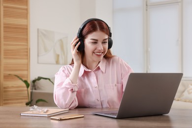 Photo of Happy woman with headphones using laptop at wooden table in room