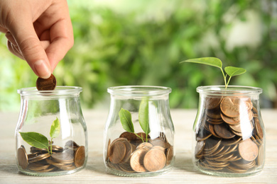 Woman putting coin into jar with green plant on white wooden table, closeup