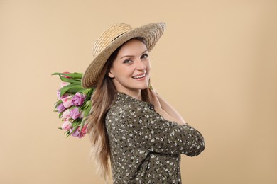 Happy young woman in straw hat holding bouquet of beautiful tulips on beige background