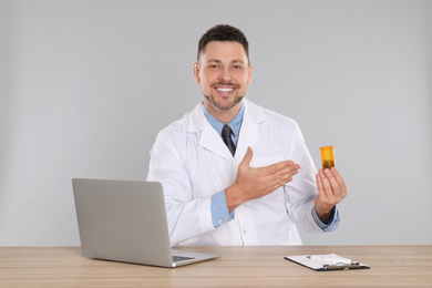 Professional pharmacist with pills and laptop at table against light grey background