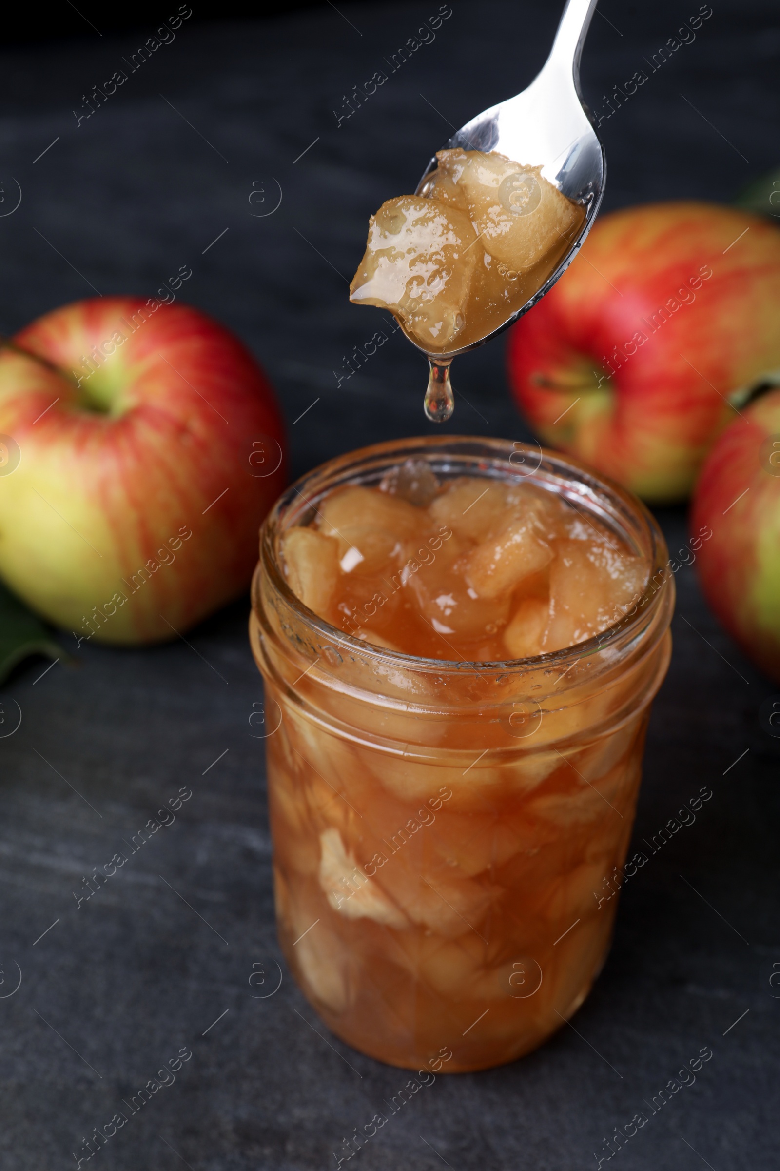 Photo of Spoon with tasty apple jam over glass jar on dark grey table