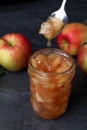 Photo of Spoon with tasty apple jam over glass jar on dark grey table