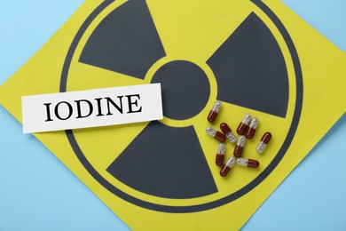 Photo of Paper note with word Iodine, pills and radiation sign on light blue background, top view