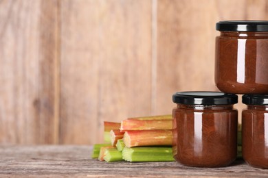 Jars of tasty rhubarb jam and stalks on wooden table, space for text