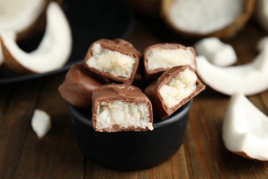 Photo of Delicious milk chocolate candy bars with coconut filling in bowl on wooden table, closeup