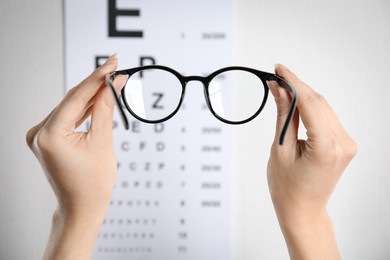 Woman holding glasses against eye chart on light background, closeup. Ophthalmologist prescription