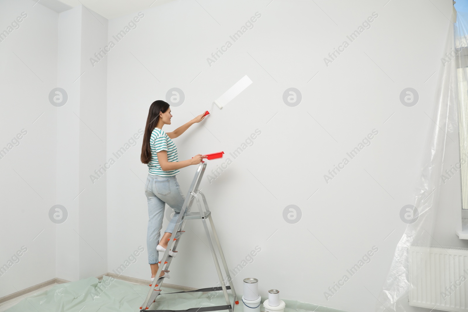 Photo of Woman standing on metallic folding ladder and painting wall indoors, space for text