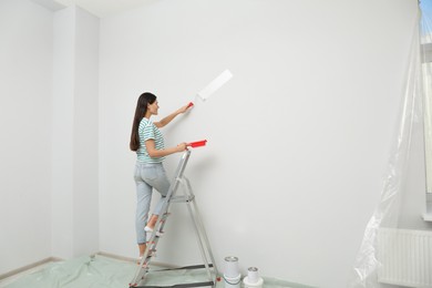 Photo of Woman standing on metallic folding ladder and painting wall indoors, space for text