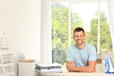 Handsome man with iron and clean laundry near board at home, space for text