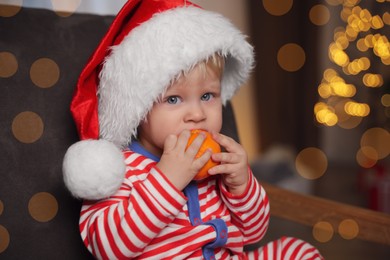 Photo of Cute little child in Santa hat with ripe tangerine in armchair at home