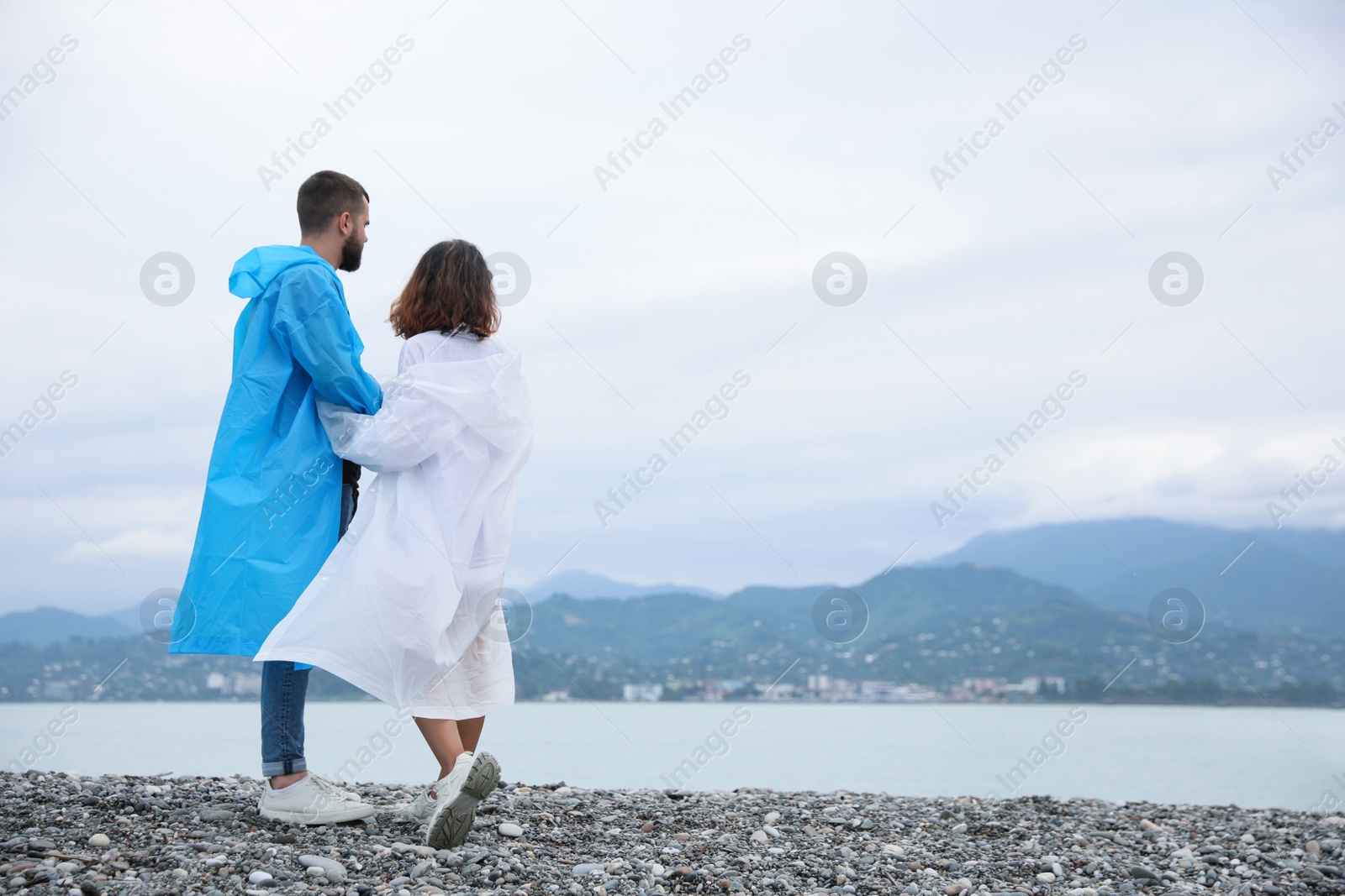 Photo of Young couple in raincoats enjoying time together under rain on beach, space for text