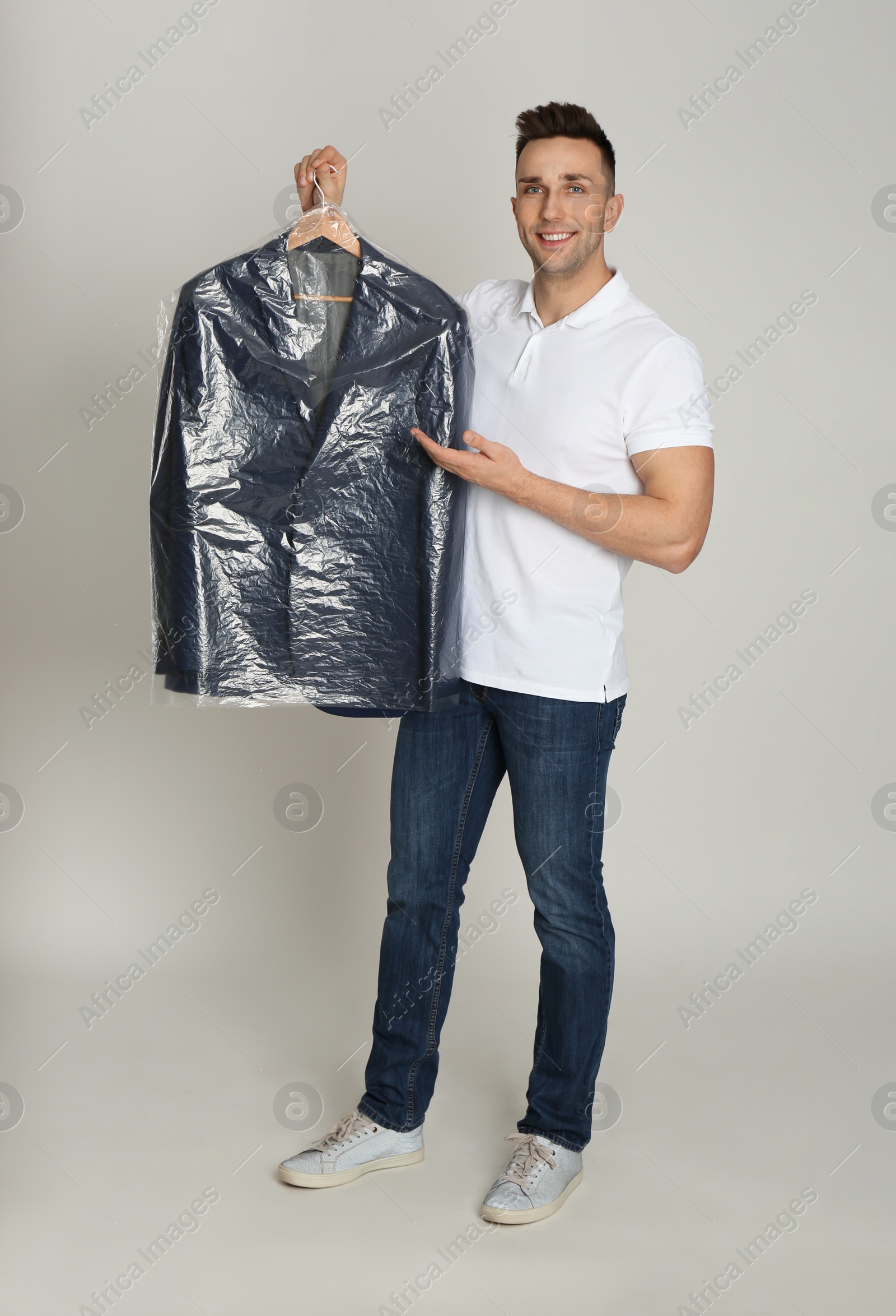 Photo of Man holding hanger with jacket in plastic bag on light grey background. Dry-cleaning service