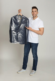 Man holding hanger with jacket in plastic bag on light grey background. Dry-cleaning service