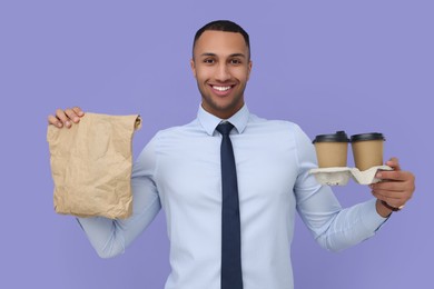 Happy young intern holding takeaway cups with hot drink and paper bag on lilac background