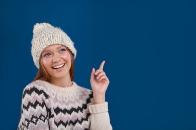 Photo of Young woman in warm sweater and hat on blue background, space for text. Winter season