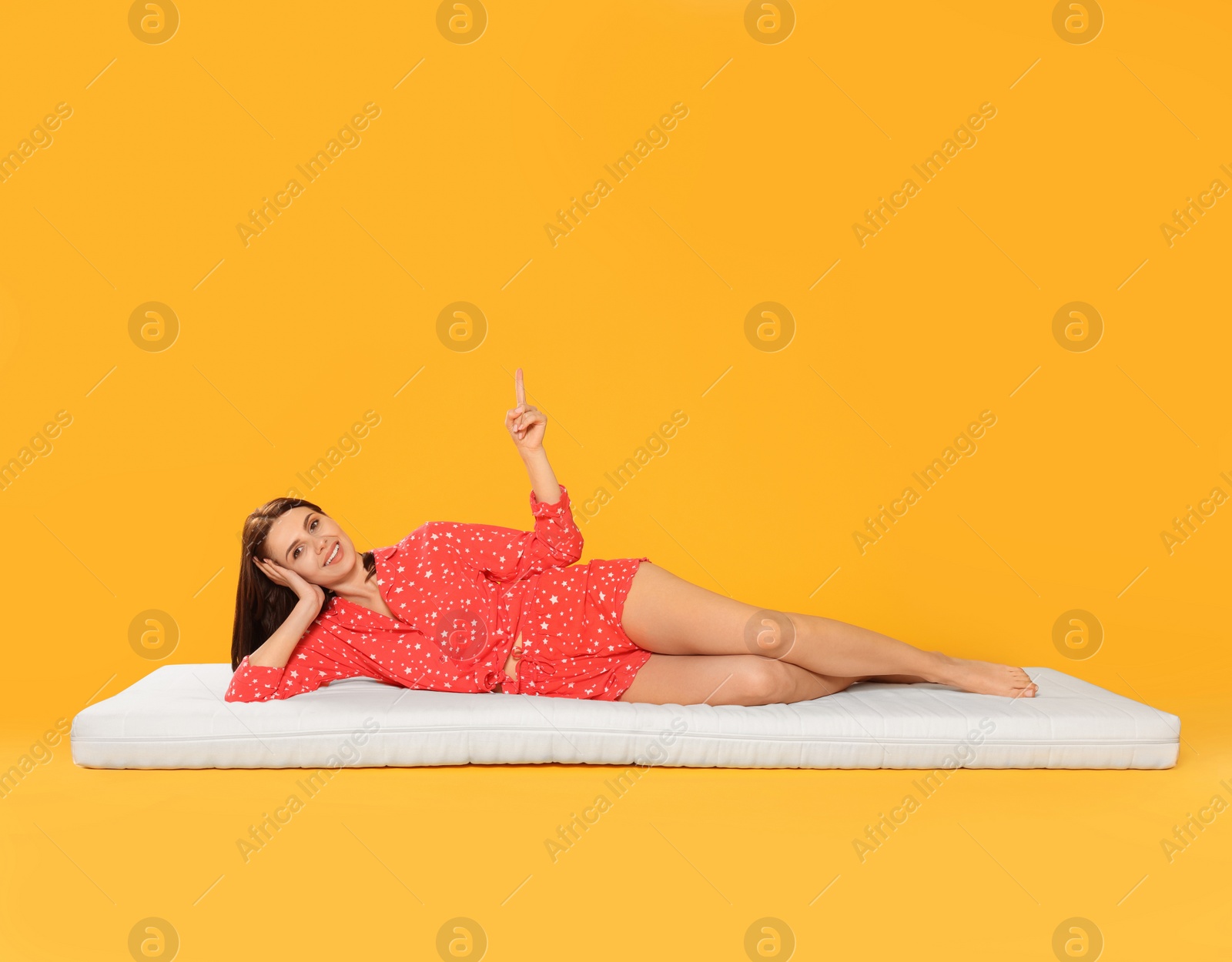 Photo of Young woman lying on soft mattress and pointing upwards against orange background