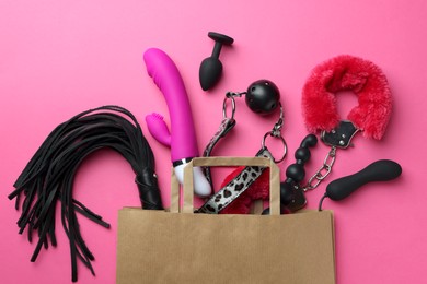 Photo of Paper shopping bag with different sex toys on pink background, flat lay