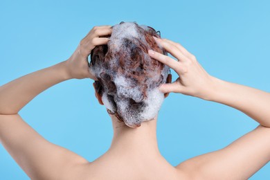 Young woman washing her hair with shampoo on light blue background, back view