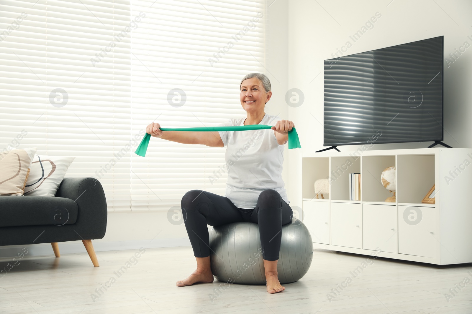 Photo of Senior woman doing exercise with elastic resistance band on fitness ball at home