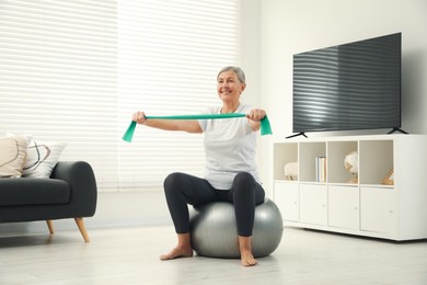 Senior woman doing exercise with elastic resistance band on fitness ball at home